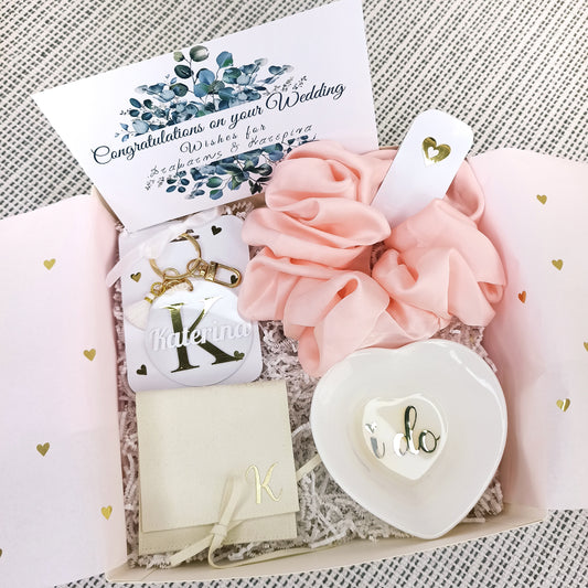 Κουτί δώρου bride to be με το όνομα της νύφης στο εξωτερικό του μέρος που περιλαμβάνει ένα κεραμικό πιατάκι κοσμημάτων που γράφει ''i do'', ένα scrunchie για τα μαλλιά σε ροζ χρώμα, μία ευχετήρια κάρτα με τα ονόματα του ζευγαριού, ένα κολιέ με το μονόγραμμά της νύφης με προσωποποιημένη βελουτέ θήκη και ένα μπρελόκ με το όνομά της.