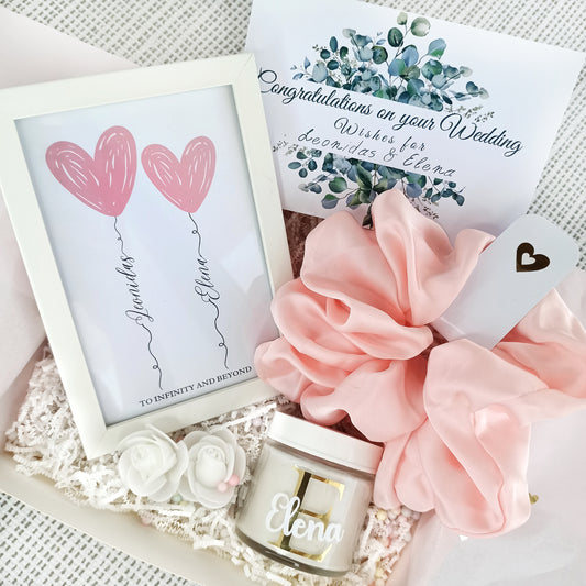 Προσωποποιημένο κουτί δώρου για νύφη με το όνομά της στο εξωτερικό του με χρυσά γράμματα και κορδέλα σε ροζ απόχρωση που περιλαμβάνει ένα καδράκι με τα ονόματα του ζευγαριού, ένα αρωματικό κερί, ένα ροζ scrunchie και μια ευχετήρια κάρτα.